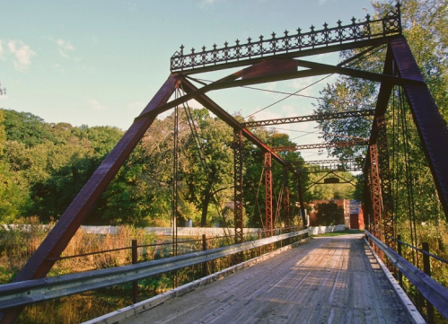 A vintage metal bridge with a wooden deck, surrounded by lush greenery and a clear blue sky.