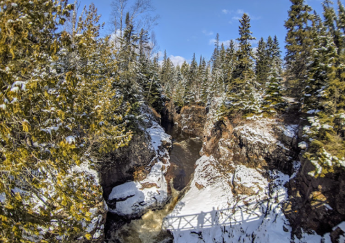A snowy landscape with a waterfall and rocky cliffs, surrounded by evergreen trees under a clear blue sky.