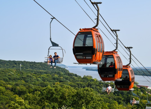 A scenic view of orange gondolas on a cable car line over lush green hills and a river in the background.