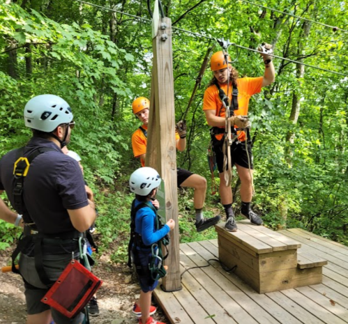 A group of people in helmets and harnesses prepares for a zip line in a wooded area.