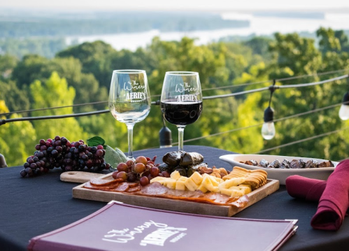 Two wine glasses beside a charcuterie board with grapes and cheese, overlooking a scenic view of greenery and water.