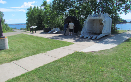 A person stands near large sculptures in a grassy area by the water, with trees and a blue sky in the background.