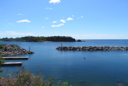 A serene coastal view featuring calm blue waters, rocky shorelines, and a clear sky with scattered clouds.