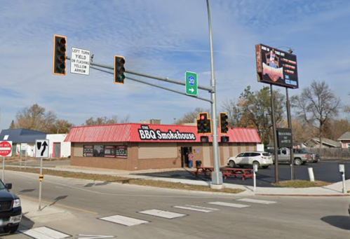 A BBQ smokehouse at an intersection with traffic lights and a large billboard nearby. Clear blue sky overhead.