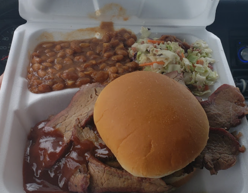 A takeout container with sliced beef in sauce, a sandwich bun, baked beans, and coleslaw.