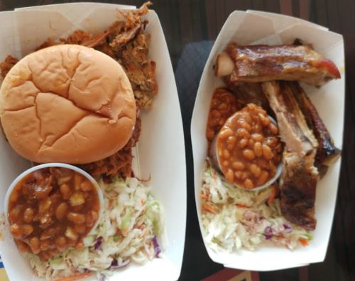 Two trays of barbecue food: one with pulled pork sandwich and baked beans, the other with ribs, baked beans, and coleslaw.