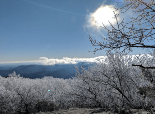 A snowy landscape with frosted trees under a bright blue sky and a shining sun, overlooking distant mountains.