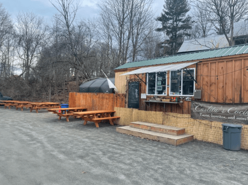 Outdoor food stand with wooden picnic tables, a menu board, and a tent in a wooded area.