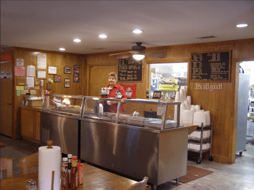 A smiling server stands behind a counter in a cozy diner, with menus and condiments visible in a warm wooden setting.