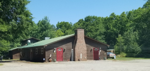 A rustic wooden building with a green roof and red doors, surrounded by trees and a gravel driveway.