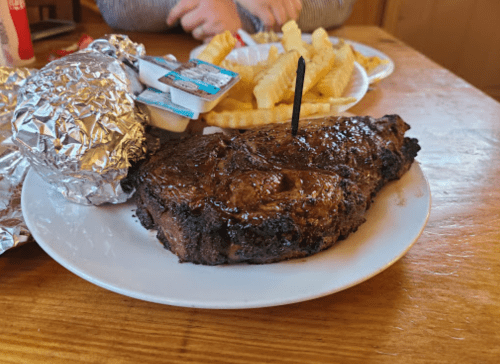 A plate with a grilled steak, a baked potato with toppings, and a side of French fries on a wooden table.