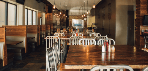 A spacious restaurant interior with wooden tables, metal chairs, and warm lighting, featuring booth seating along the walls.