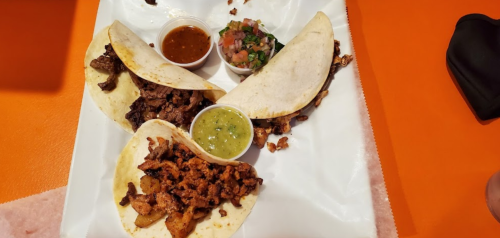 Three tacos filled with different meats, accompanied by salsa, pico de gallo, and green sauce on a white plate.