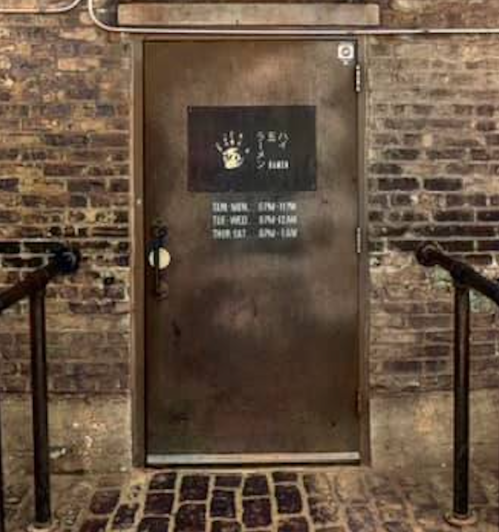 A metal door with a sign displaying bakery hours, set against a brick wall and surrounded by metal railings.