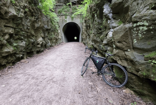 A bike rests on a dirt path beside a rocky tunnel entrance surrounded by greenery.