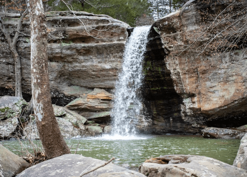 A serene waterfall cascades over rocky cliffs into a calm green pool, surrounded by trees and natural stone formations.