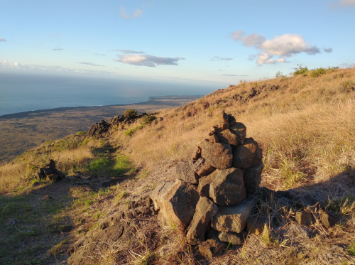 A rocky cairn on a grassy hillside overlooking a coastal landscape under a clear blue sky.