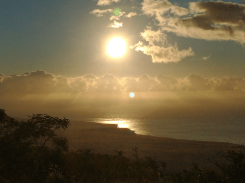 A golden sunset over a calm ocean, with clouds reflecting the warm light and a distant shoreline visible.