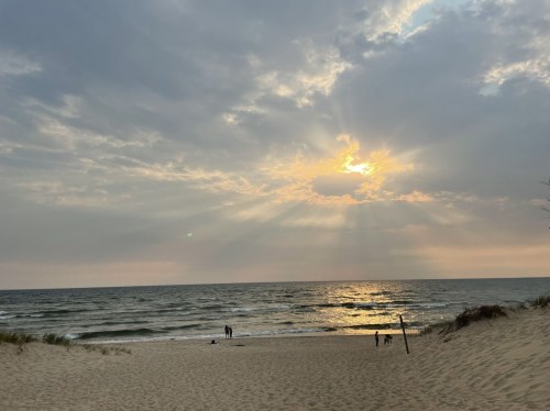 A serene beach scene at sunset, with gentle waves and people walking along the shore under a cloudy sky.
