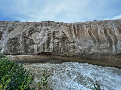 A rugged cliffside with textured rock formations above a flowing river under a cloudy sky.