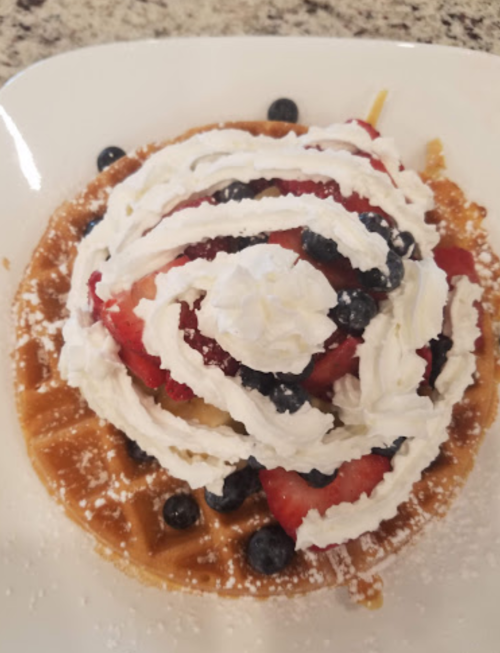 A waffle topped with strawberries, blueberries, and whipped cream, garnished with powdered sugar on a white plate.