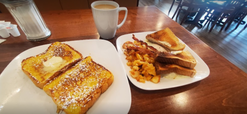 A plate of French toast with butter and powdered sugar, alongside a plate of toast, bacon, and hash browns, with coffee.