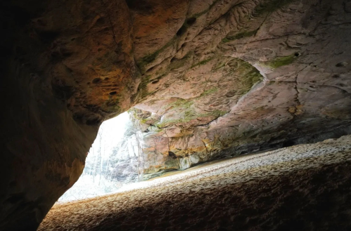 A rocky cave entrance with light streaming in, revealing a sandy floor and textured stone walls.