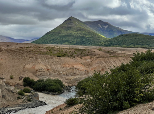 A scenic landscape featuring a mountain, river, and lush greenery under a cloudy sky.