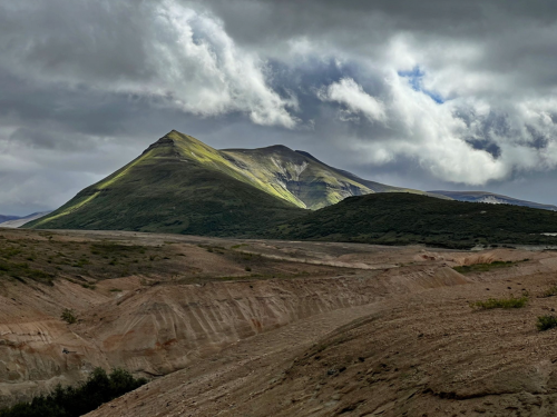 A dramatic mountain peak under a cloudy sky, with sunlight illuminating its slopes and a barren landscape in the foreground.