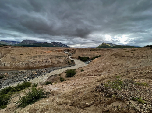 A rugged landscape with a winding river, rocky terrain, and dramatic, cloudy skies in the background.