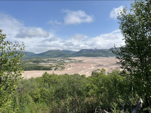 A panoramic view of a dry, sandy landscape surrounded by green trees and distant mountains under a cloudy sky.