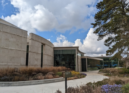 Modern building with a curved facade, surrounded by landscaped gardens and under a partly cloudy sky.