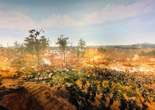A detailed diorama depicting a chaotic battle scene with soldiers, cannons, and a burning building amidst a forested landscape.