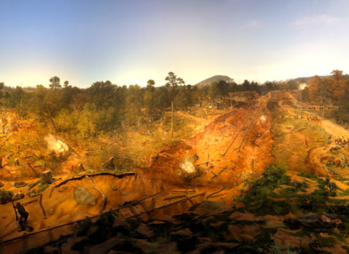 A panoramic view of a battlefield scene with smoke, trees, and soldiers amidst a rugged landscape.