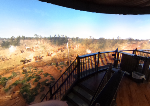 A panoramic view from a balcony overlooking a dusty landscape with trees and distant figures, possibly on horseback.