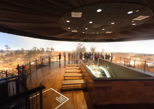 A panoramic view from an observation deck with people standing near a railing, overlooking a scenic landscape.