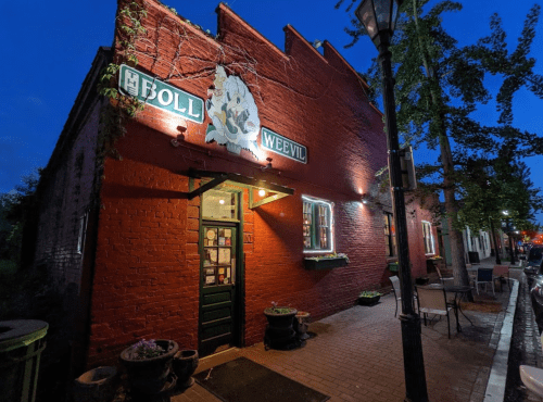 A red brick building with a sign reading "The Boll Weevil," illuminated at dusk, surrounded by trees and outdoor seating.
