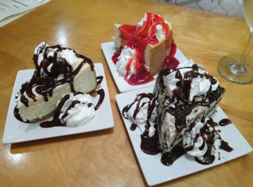 Three slices of cheesecake topped with whipped cream and chocolate sauce, one with strawberry topping, on white plates.