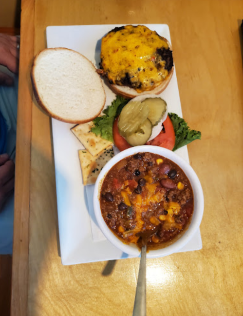 A plate featuring a cheeseburger with pickles, lettuce, tomato, crackers, and a bowl of chili topped with cheese.
