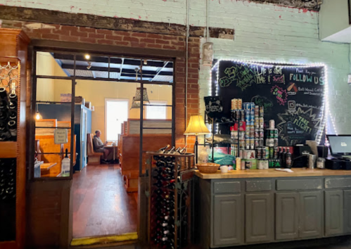 A cozy café interior with a counter displaying drinks and a chalkboard, leading to a seating area in the background.