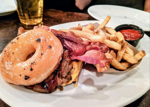 A glazed donut sandwich with bacon and pulled pork, served with a side of fries and ketchup.