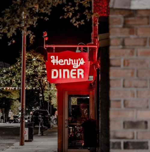 A glowing red sign for "Henry's Diner" at night, framed by brick and trees with festive lights in the background.