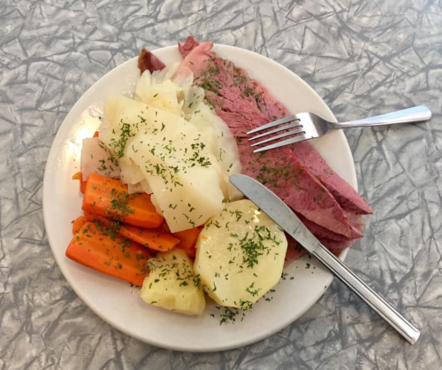 A plate of corned beef with carrots, potatoes, and cabbage, garnished with herbs, on a textured table surface.