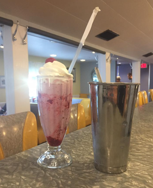 A tall glass of strawberry milkshake topped with whipped cream and a cherry, next to a metal cup.