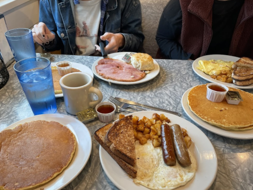 A table with various breakfast dishes, including pancakes, eggs, sausage, ham, and toast, with drinks in blue glasses.