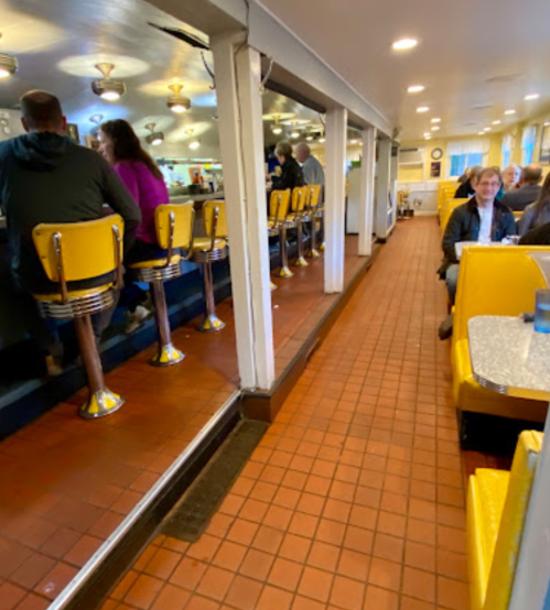 A retro diner interior with yellow stools at the counter and booths along the wall, featuring tiled flooring.