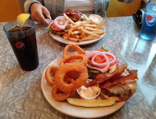 A table with two plates of burgers topped with bacon, fries, onion rings, and drinks.