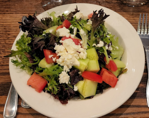 A fresh salad with mixed greens, diced tomatoes, cucumber, and crumbled feta cheese in a white bowl.