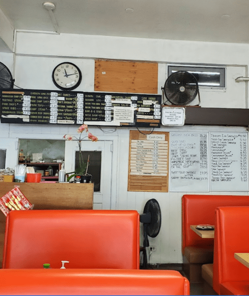Interior of a casual restaurant featuring orange seating, a menu board, clock, and wall-mounted fans.