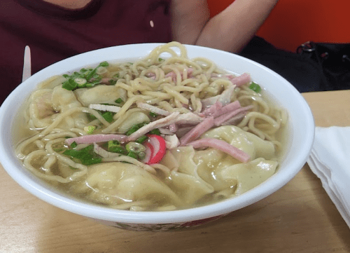 A bowl of noodle soup with dumplings, sliced meat, and green onions, placed on a wooden table.